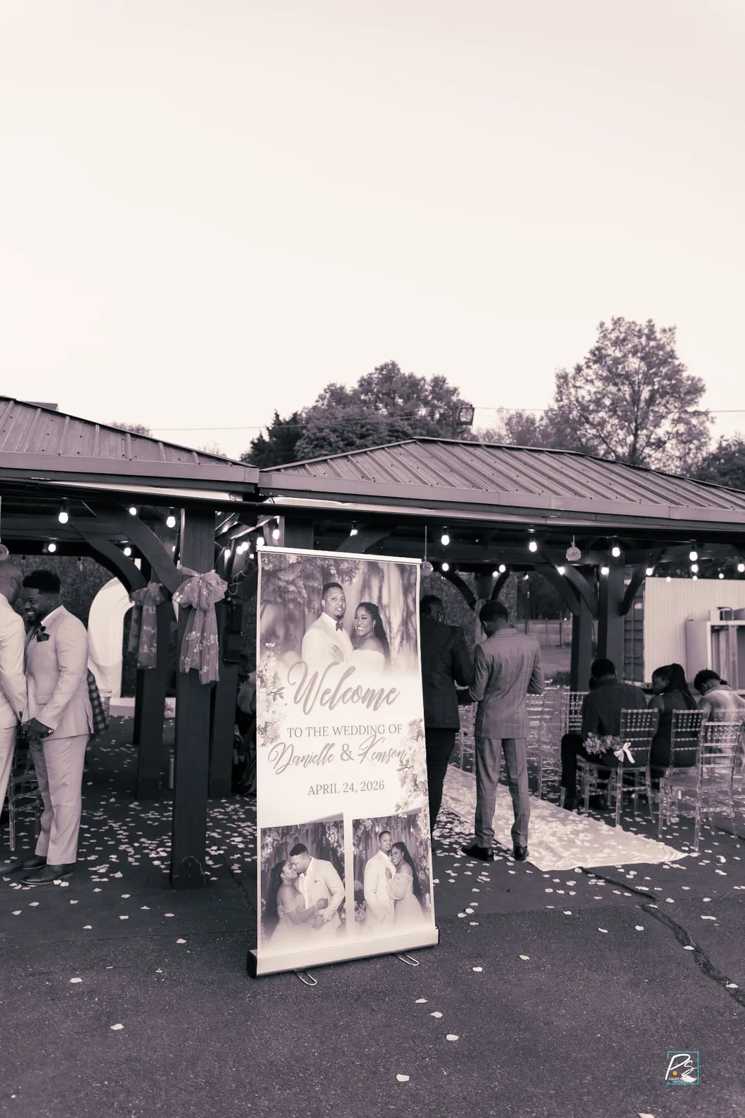 Outdoor wedding entrance with a welcome sign banner, petal-scattered aisle and clear chiavari chairs under a string-lit pavilion
