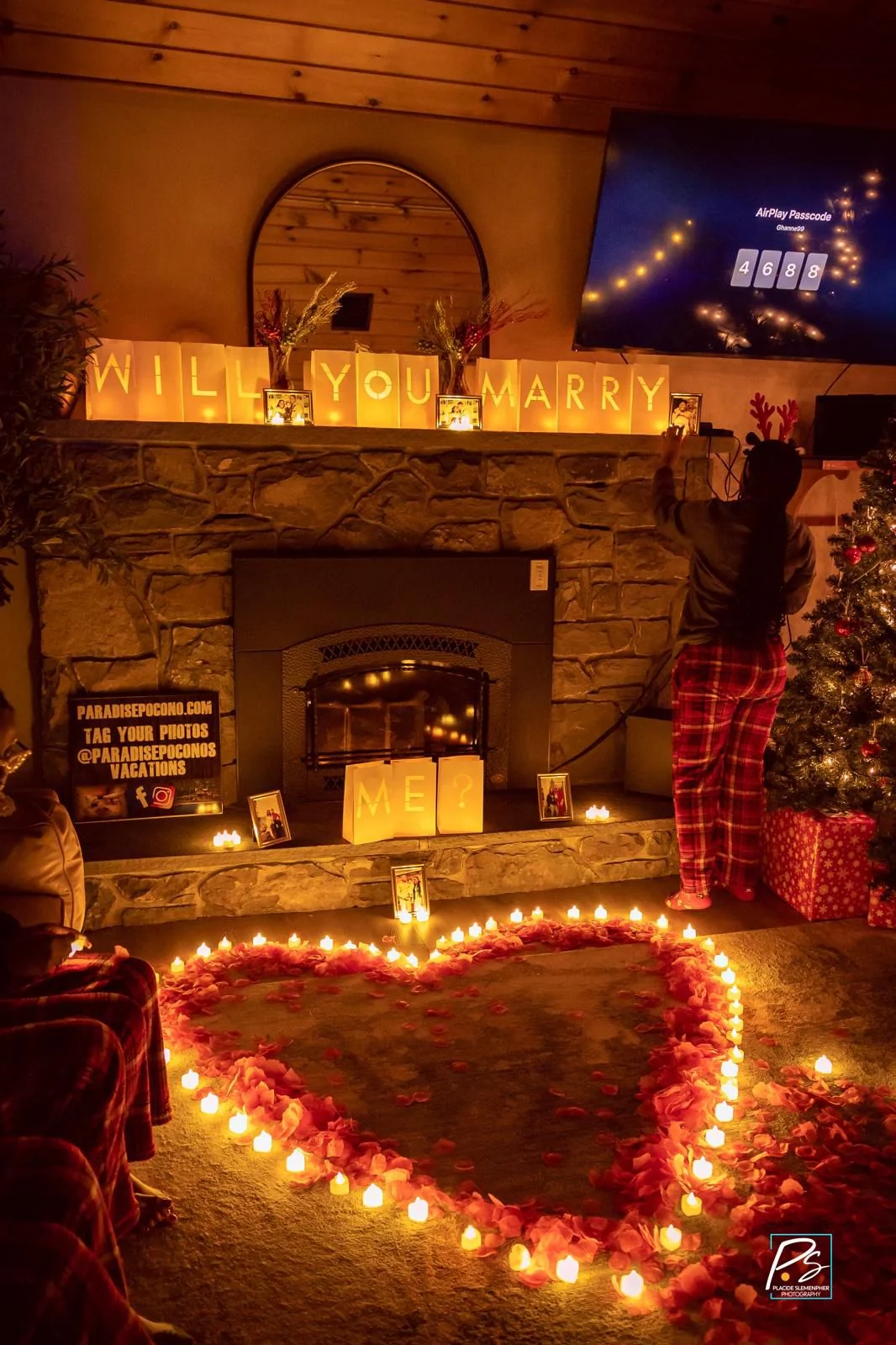 Candlelit proposal setup with illuminated will you marry me lettering on a stone fireplace and a rose petal heart on the floor