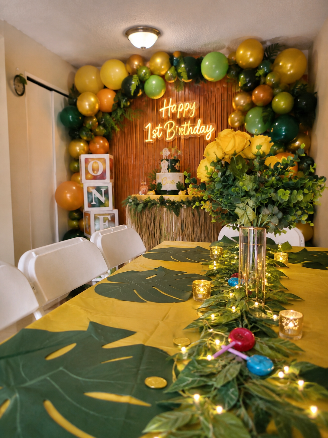 Jungle themed first birthday setup with green and gold balloon arch, Happy 1st Birthday neon sign, cake table and a tropical leaf tablescape with candles
