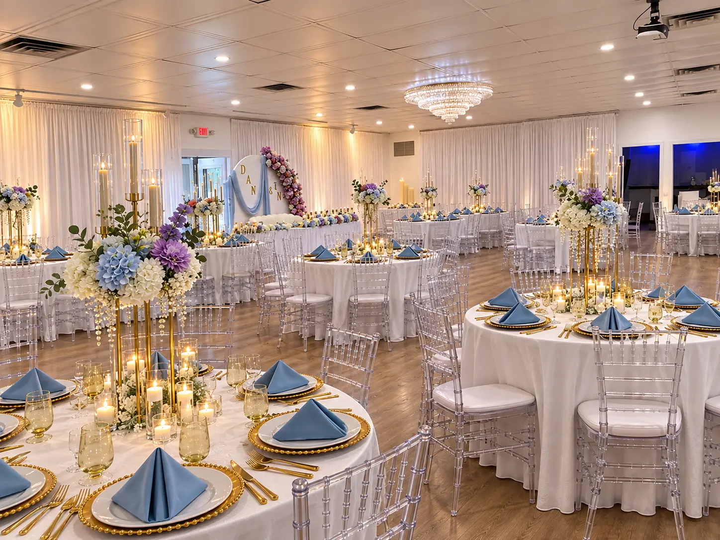 Wedding reception ballroom with round tables, dusty blue napkins, gold chargers, tall floral centerpieces and clear chiavari chairs