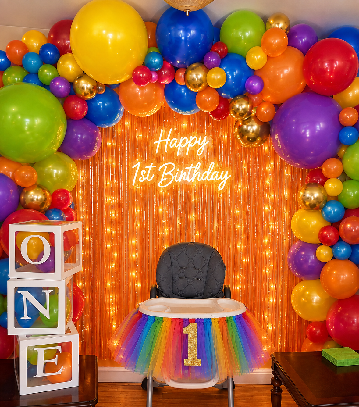 Colorful first birthday setup with a rainbow balloon garland, gold fringe backdrop, Happy 1st Birthday neon sign, ONE blocks and a tutu-decorated high chair