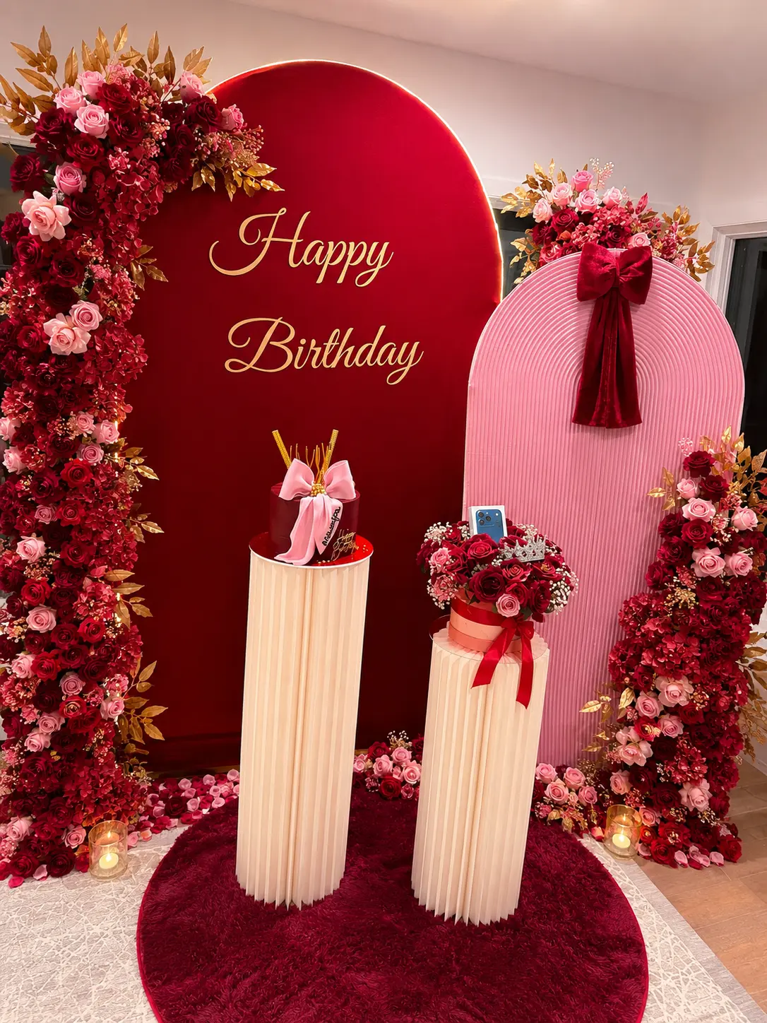 Red and pink birthday arch backdrops framed with roses, gold foliage, pedestals and a cake display