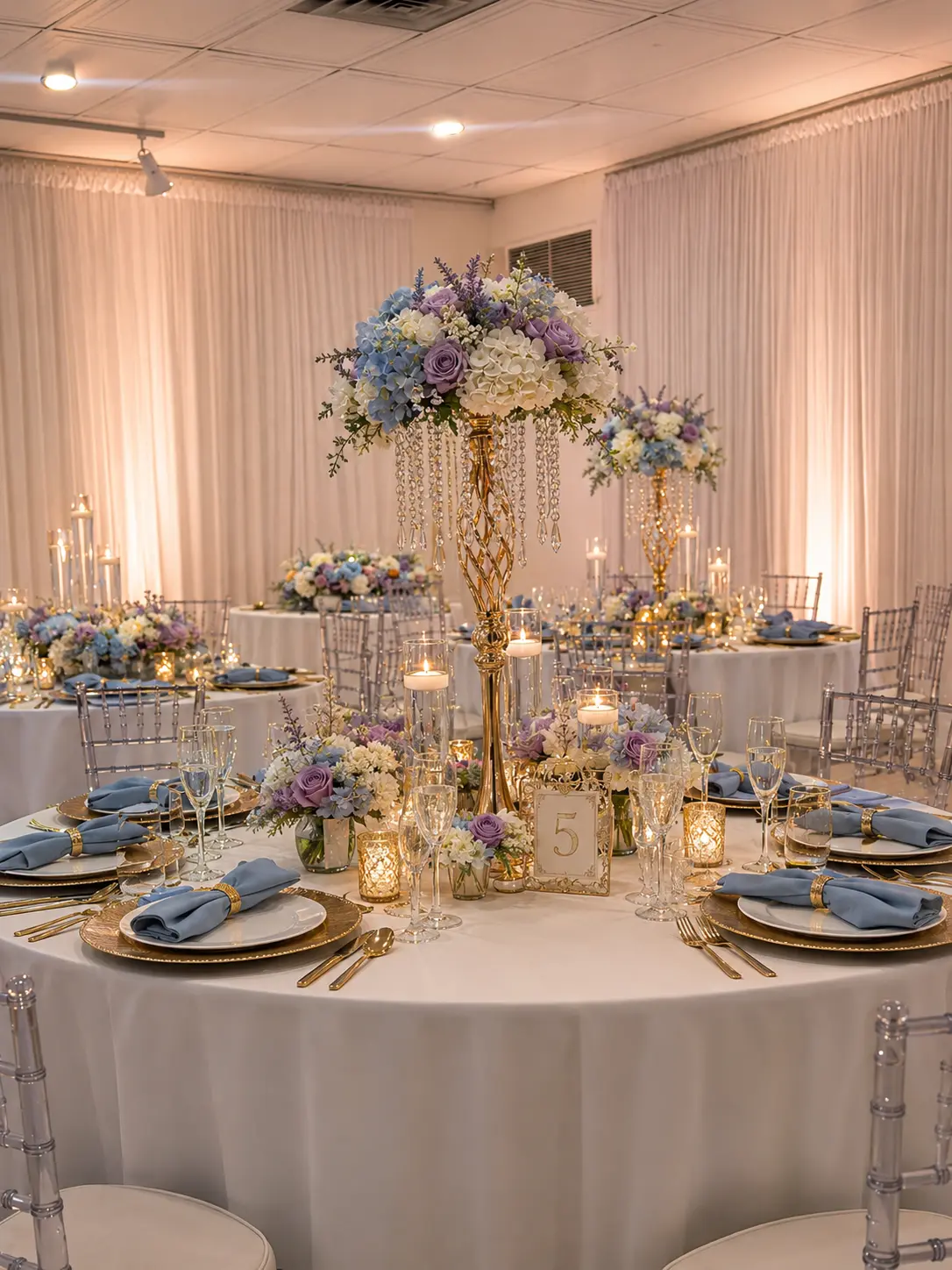 Round guest table with a tall crystal draped floral centerpiece, gold chargers, blue napkins and votive candles