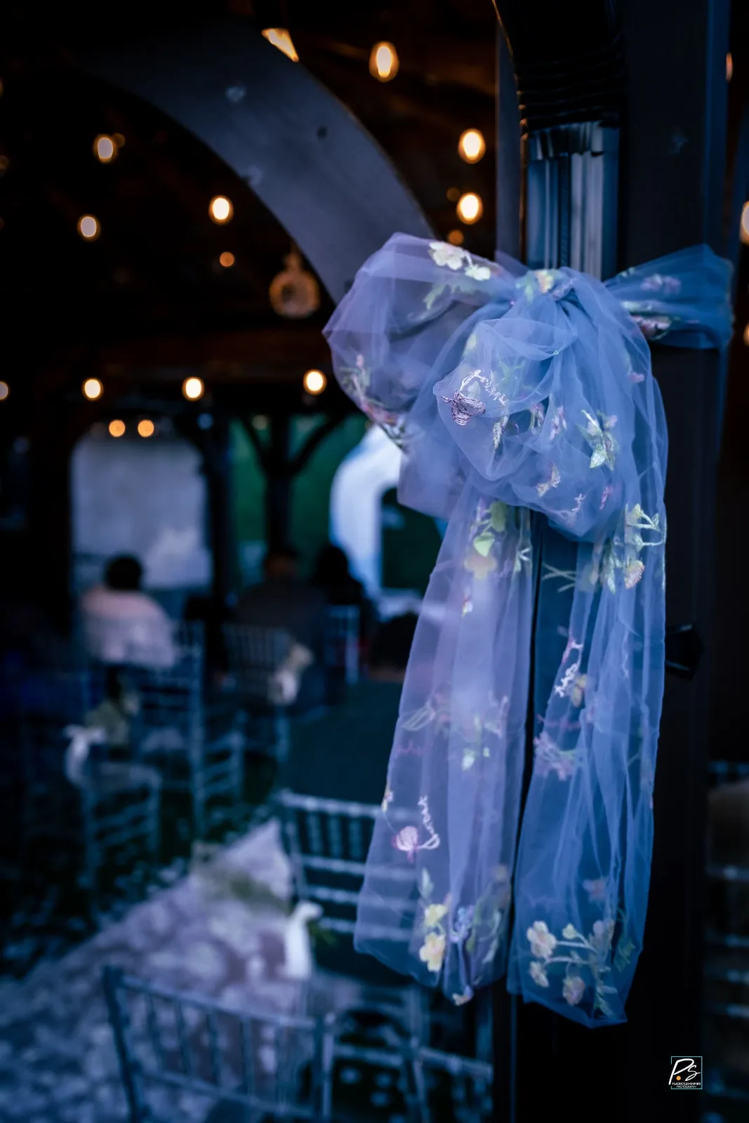 Ceremony aisle detail with an embroidered floral tulle bow tied to a post, clear chiavari chairs and warm string lights