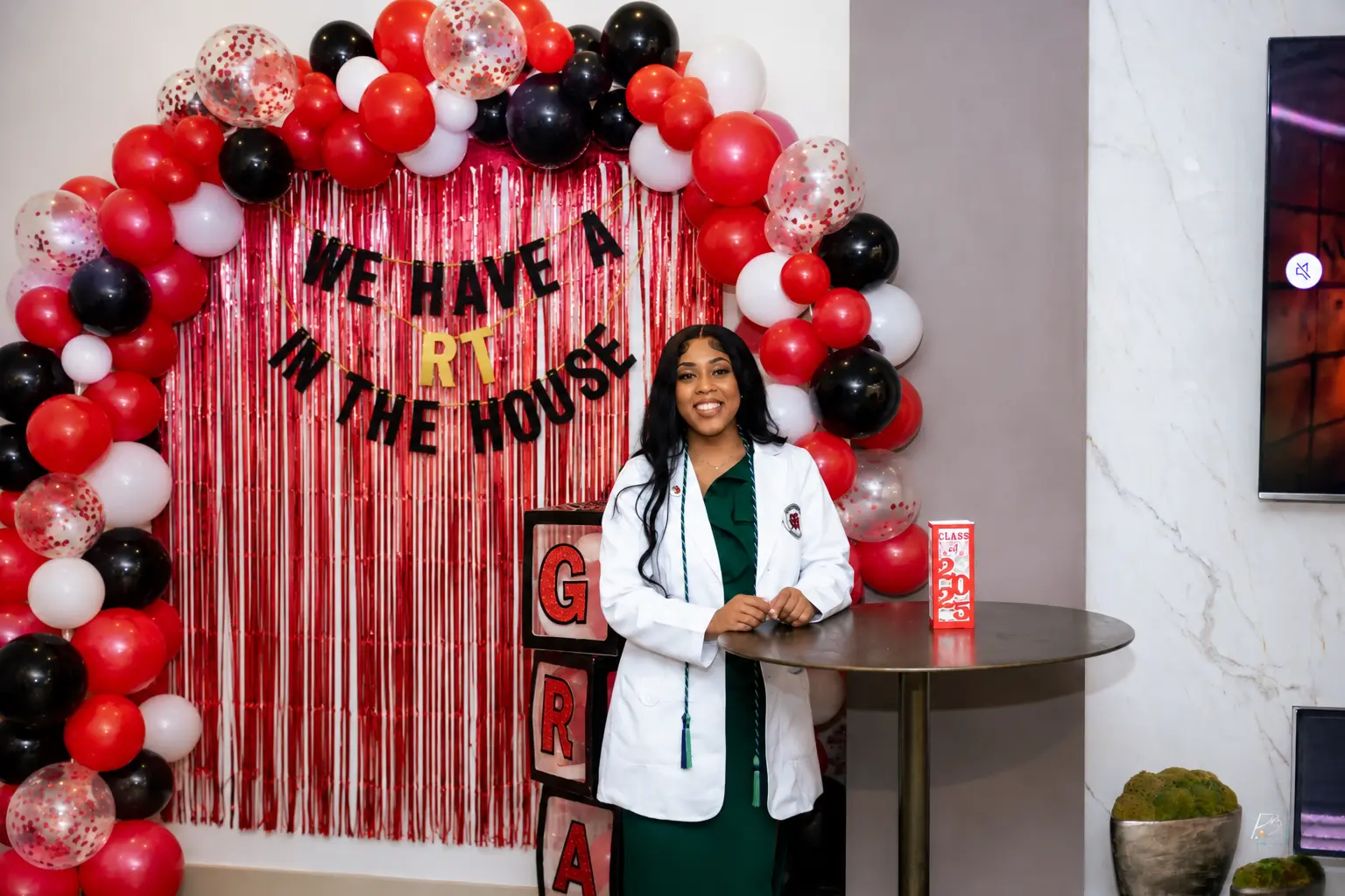 Graduation setup with a red, black and white balloon arch, fringe backdrop and celebration lettering