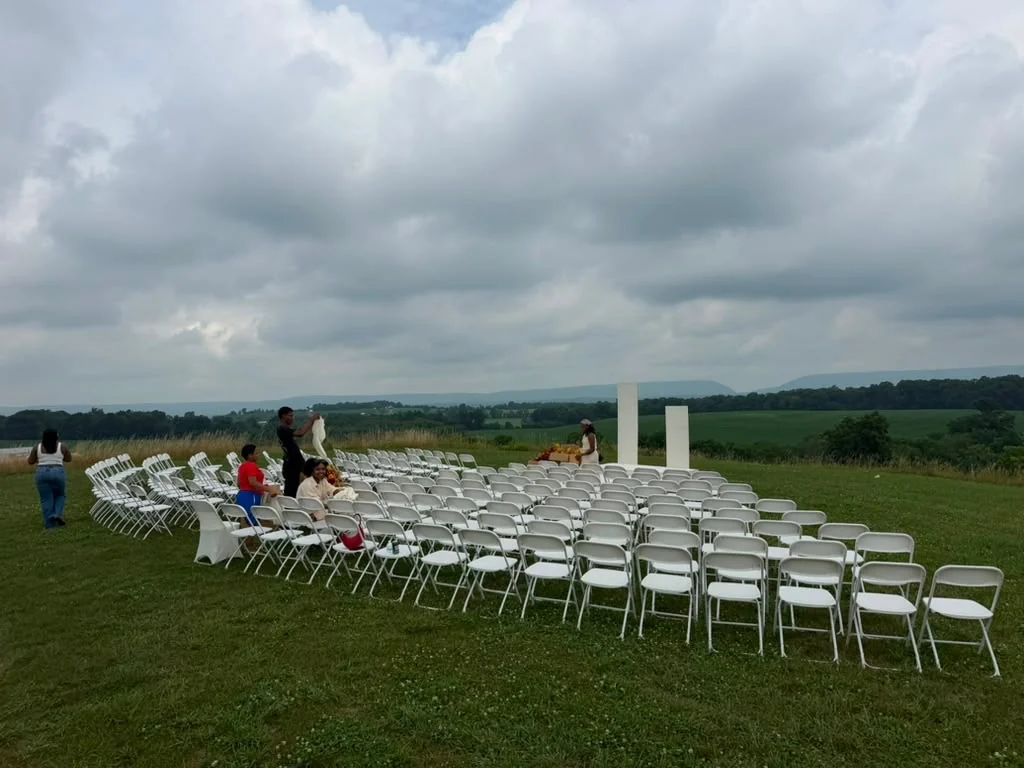 Outdoor ceremony setup with rows of white folding chairs and an aisle arch overlooking open countryside