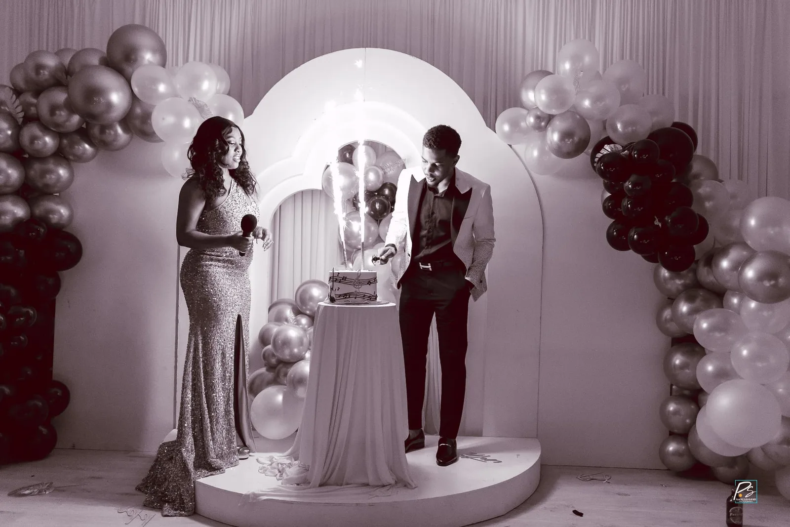 Celebration setup with an arched backdrop, balloon garlands, draped cake table and sparkler candles on a round platform